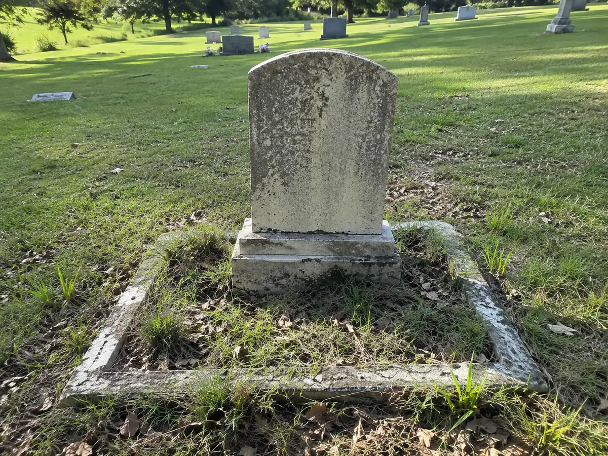 A weathered, lichen-stained headstone with an overgrown, weedy stone border before cleaning.